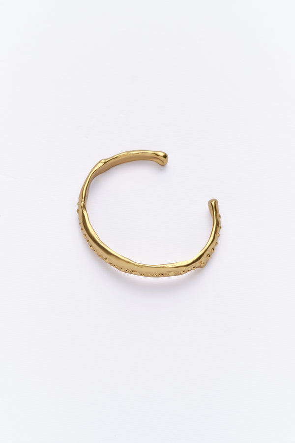 A person's forearm wearing a gold-colored cuff bracelet with a vintage look, resting on a chair.