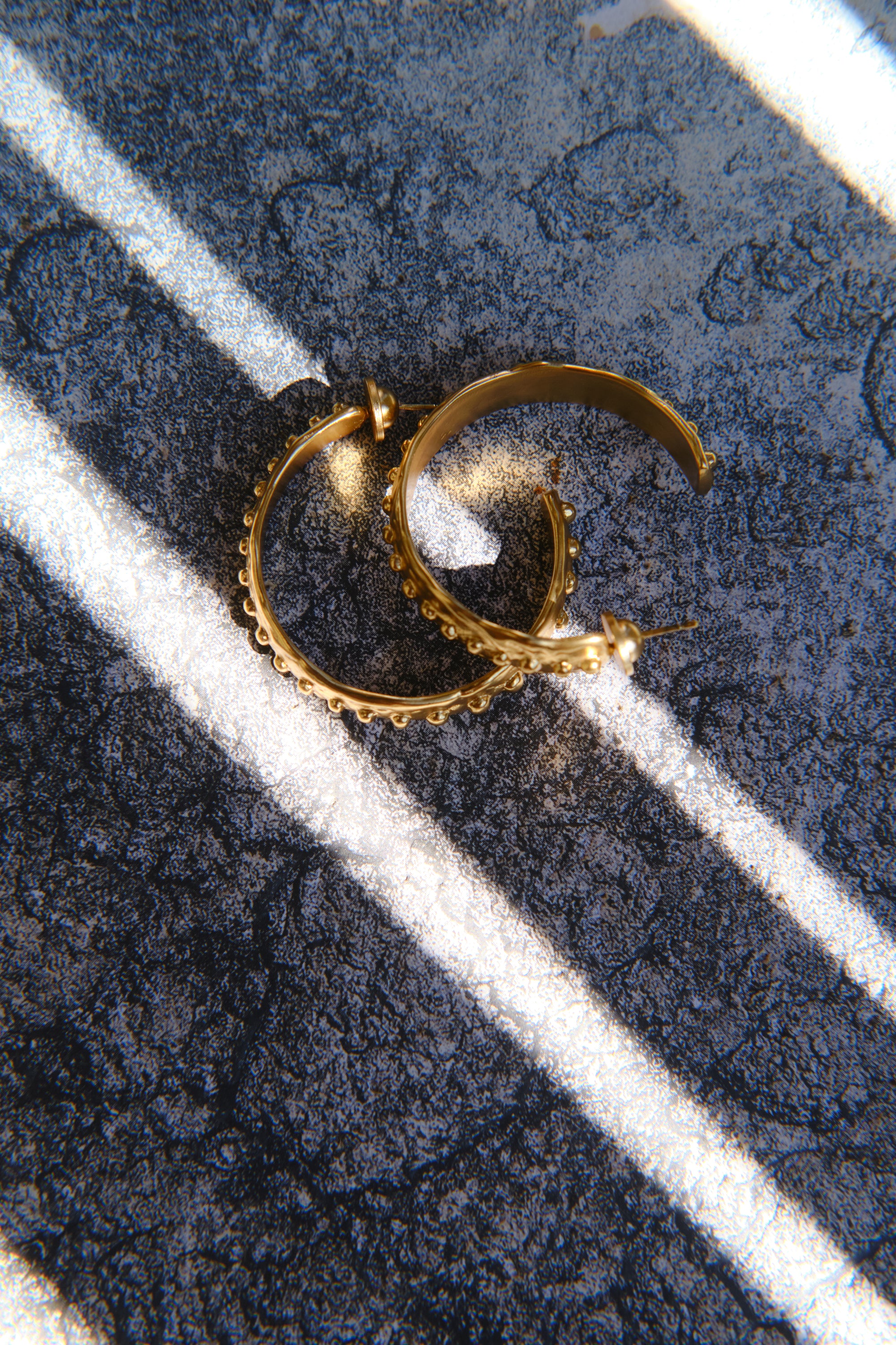 Gold textured hoop earrings styled on stone surface with light shadow