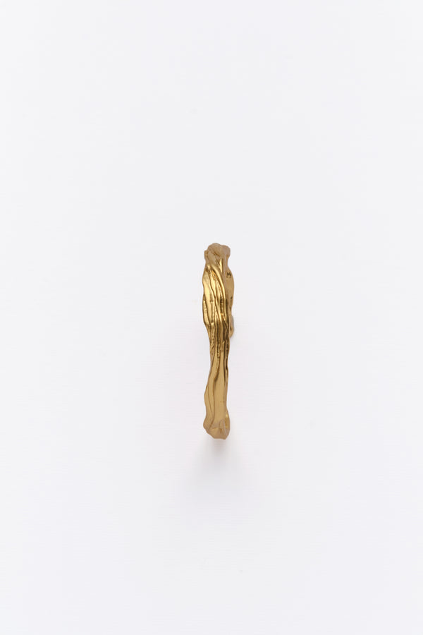 A gold-colored cuff bracelet with a textured design, resembling eroded soil layers, displayed against a white background.