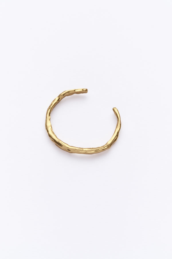A gold-colored cuff bracelet with a textured design, resembling eroded soil layers, displayed against a white background.