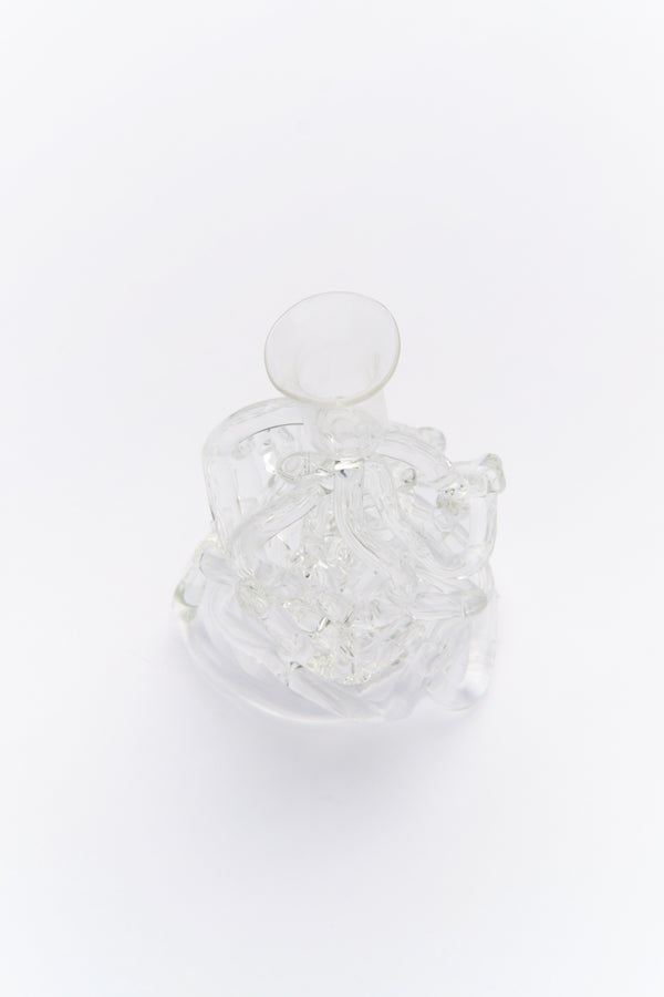 A clear glass candle holder with a sculpted design, resembling the movement of water, displayed with a lit candle on a white surface.