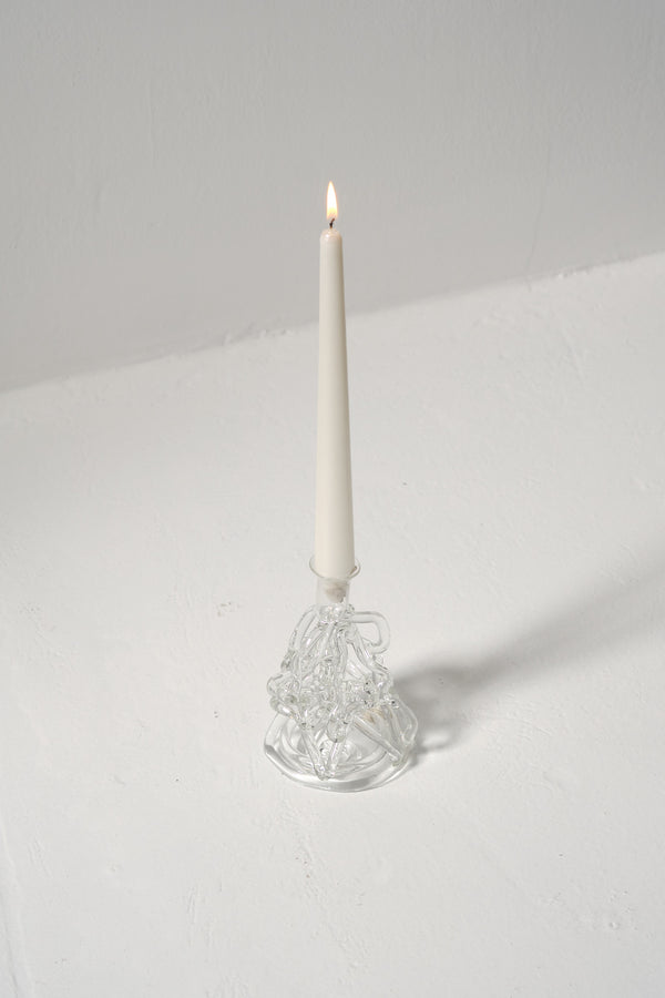 A clear glass candle holder with a sculpted design, resembling the movement of water, displayed with a lit candle on a white surface.