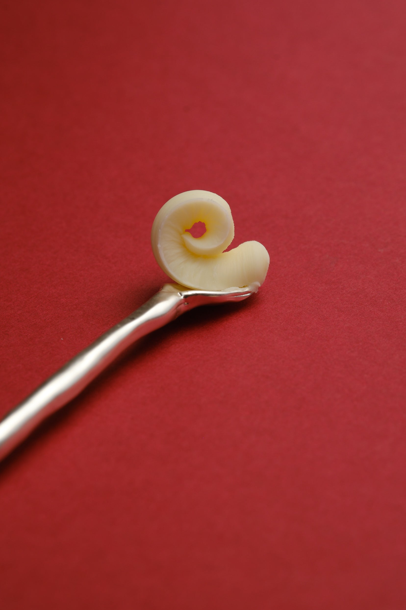 Close-up detail of sculptural silver stirring stick with butter curl on red background