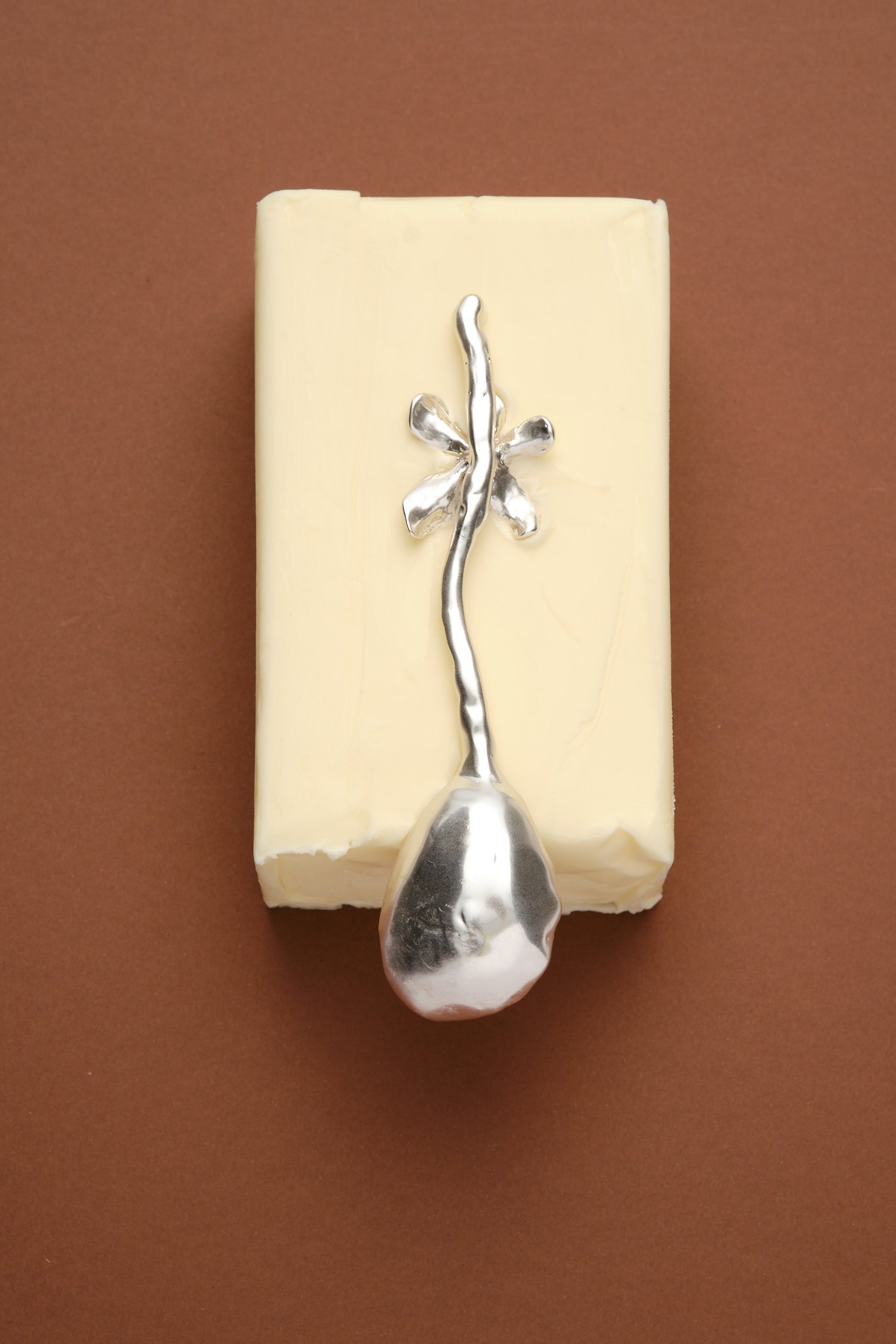 Artistic sculptural silver spoon with organic floral handle placed on butter block on brown background