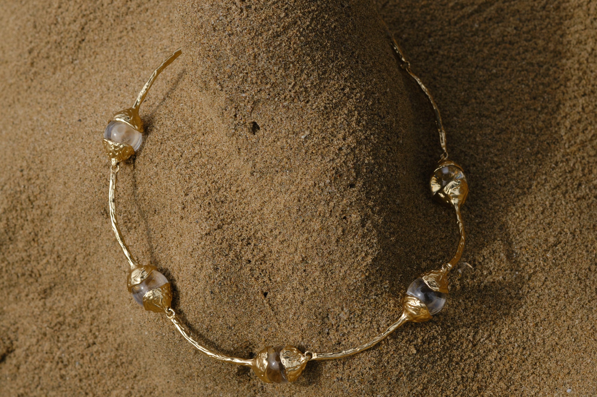 Sculptural gold necklace with organic pearl details styled on textured sand background