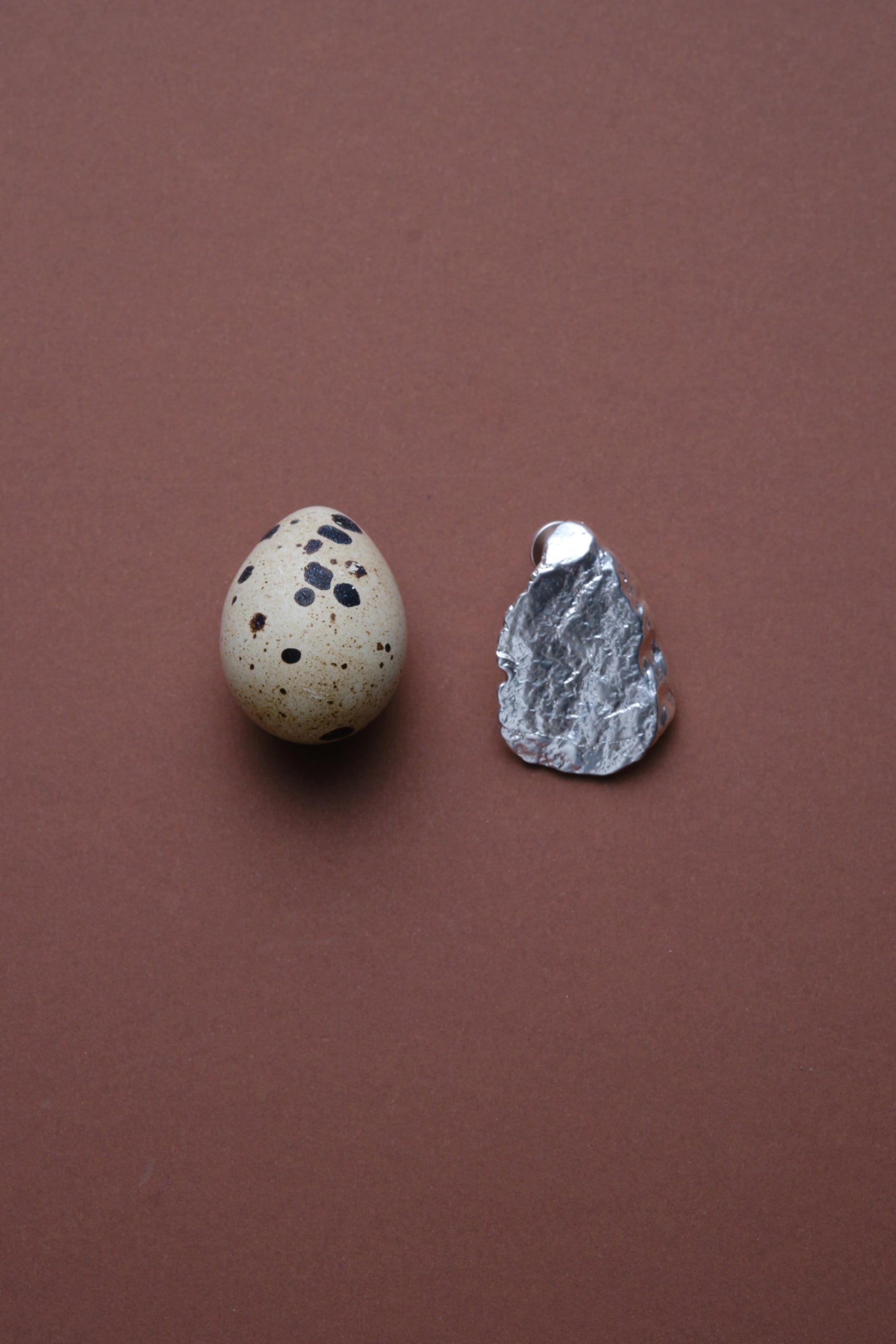 Sculptural textured silver jewelry piece displayed next to speckled egg on brown background