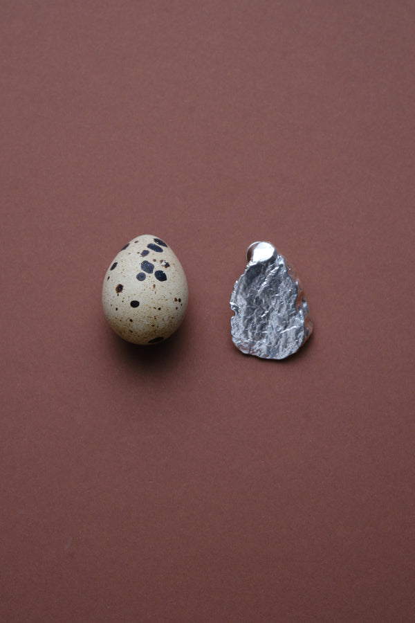 Sculptural textured silver jewelry piece displayed next to speckled egg on brown background