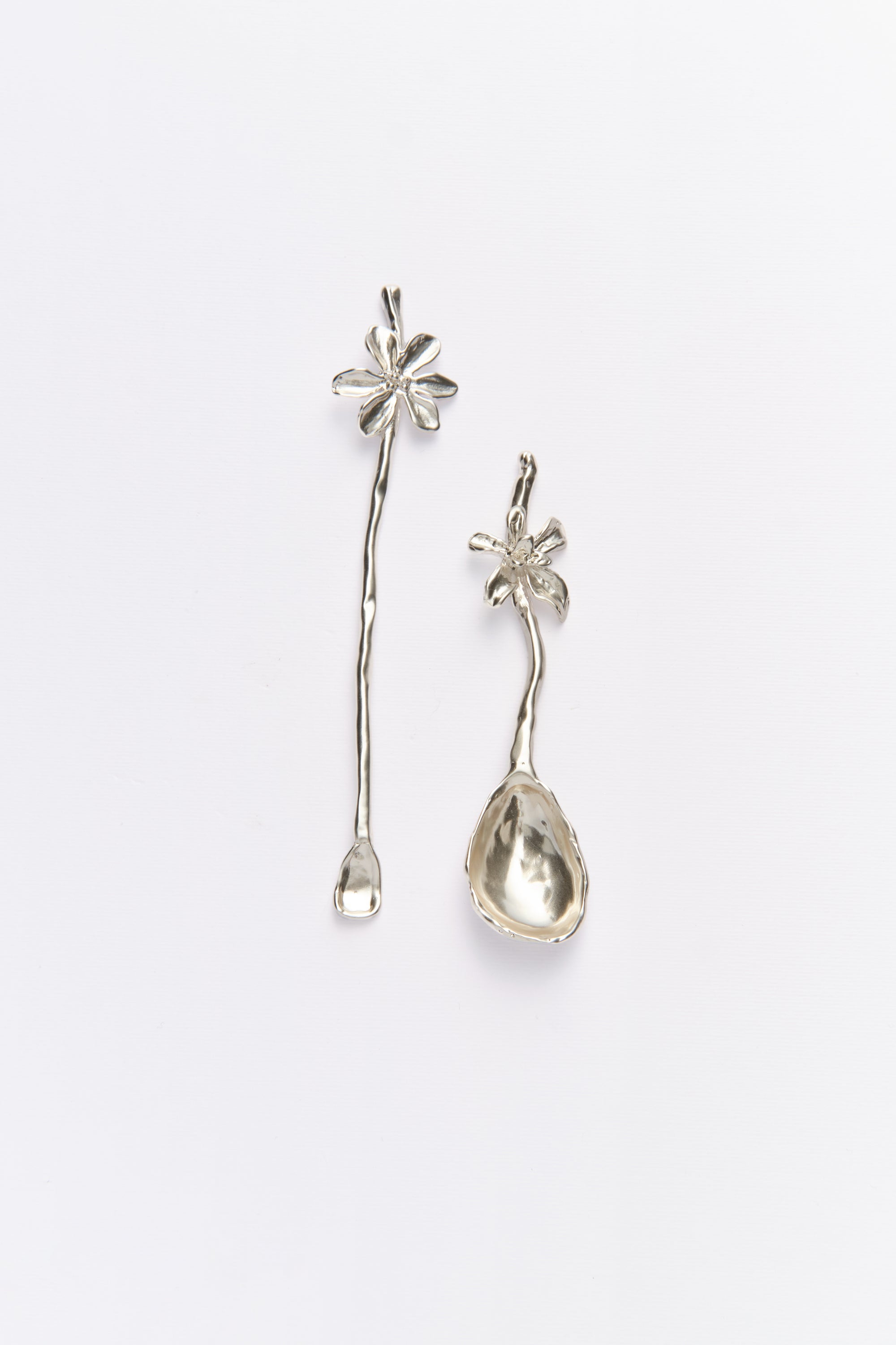 Two silver flatware items, one designed as a spoon and the other as a stirring stick, both with organic metal shapes and floral elements.